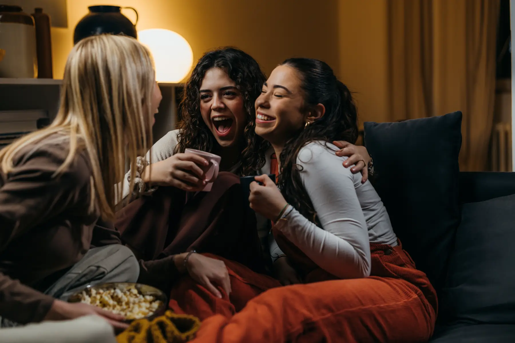 Three friends on the sofa, laughing together, popcorn and a blanket.