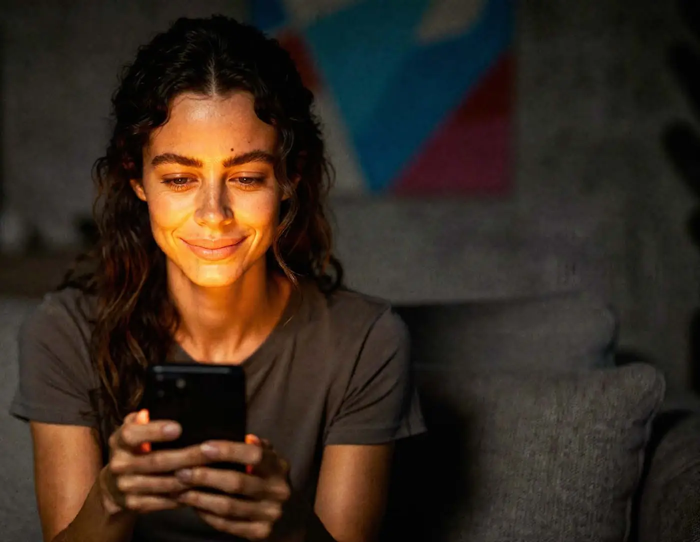 A woman on the sofa at night, smiling, phone in hand, the TV on behind her.